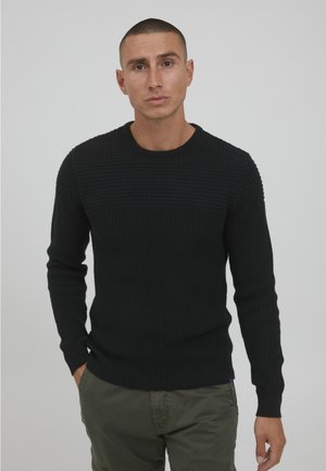 Maglione nero a maglia con un motivo testurizzato sulla parte superiore del petto. Presenta un colletto rotondo e maniche lunghe, abbinato a pantaloni verdi.