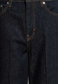 Jeans in denim blu scuro con cuciture arancioni, cinque tasche e dettagli in metallo argentato. Presentano un design classico a gamba dritta e una chiusura con zip.