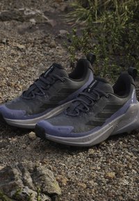 Šedé atletické boty s texturovaným povrchem, s logem GORE-TEX, šněrovacím designem a podrážkou s fialovými akcenty, umístěné na štěrkovém pozadí.