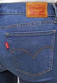 Vaqueros de mezclilla azul con un parche de cuero marrón que presenta el texto "Levi Strauss & Co." y una etiqueta roja en el bolsillo trasero, caracterizados por su costura clásica.