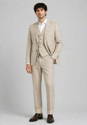 Homme debout, une main dans la poche, portant un costume beige, un gilet, une chemise blanche et des chaussures habillées marron foncé sur un fond gris uni.