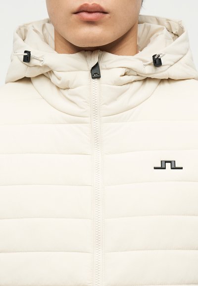 Μπεζ μπουφάν με οριζόντια ραφή, εμπρόσθιο φερμουάρ, κουκούλα με ρυθμιζόμενα κορδόνια και μαύρο λογότυπο στο στήθος.
