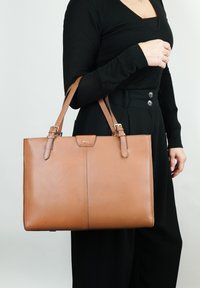 Bruine leren schoudertas met dubbele handvatten, een gladde textuur en een gestructureerde rechthoekige vorm. Voorzien van een eenvoudige voorklepsluiting en subtiele branding.