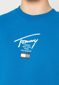 Blaues Baumwoll-T-Shirt mit Rundhalsausschnitt. Verfügt über ein weißes Logo und den Text "Tommy" mit Städtenamen sowie einem rechteckigen Aufdruck "TOMMY JEANS".