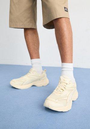Benedenbenen met beige shorts, witte sportsokken en crèmekleurige chunky sneakers staand op een blauwe vloer tegen een witte muur.