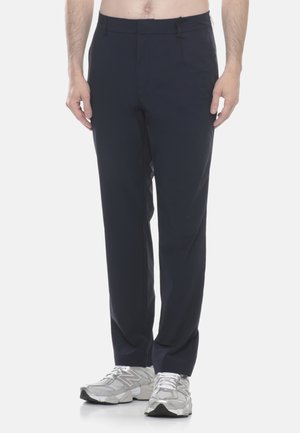 Uomo che indossa pantaloni dritti blu navy e sneakers bianche New Balance, in piedi davanti a uno sfondo bianco uniforme.