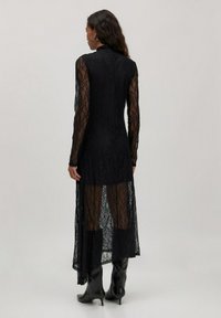 Zwarte lace jurk met lange mouwen, hoge hals, doorzichtige onderkant en een vloeiende design. G gedragen met kniehoge zwarte laarzen met hak.
