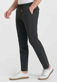 Man in slim-fit donkergrijze casual broek met een trekkoord in de taille en witte sneakers, staande met één hand in de zak.