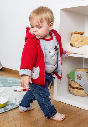 Rote Zip-Up-Hoodie mit Tiergesicht-Details über einem grauen Shirt und blauen Jeans. Kind steht in der Nähe eines Regals mit Plüschtieren.