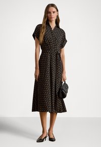 Vestido preto com padrão geométrico em amarelo, mangas curtas, cintura marcada e saia fluida. Modelo segurando uma pequena mala preta.