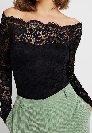 Woman wearing a black off-the-shoulder lace top with floral patterns and light green high-waisted corduroy pants with hands in pockets.