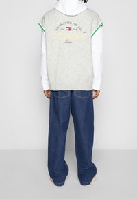 Grauer gestrickter ärmelloser Pullover mit Tommy Jeans-Branding, über einem weißen Kapuzenpullover getragen, kombiniert mit lockeren blauen Denim-Jeans. Grüne Akzente am Pullover.