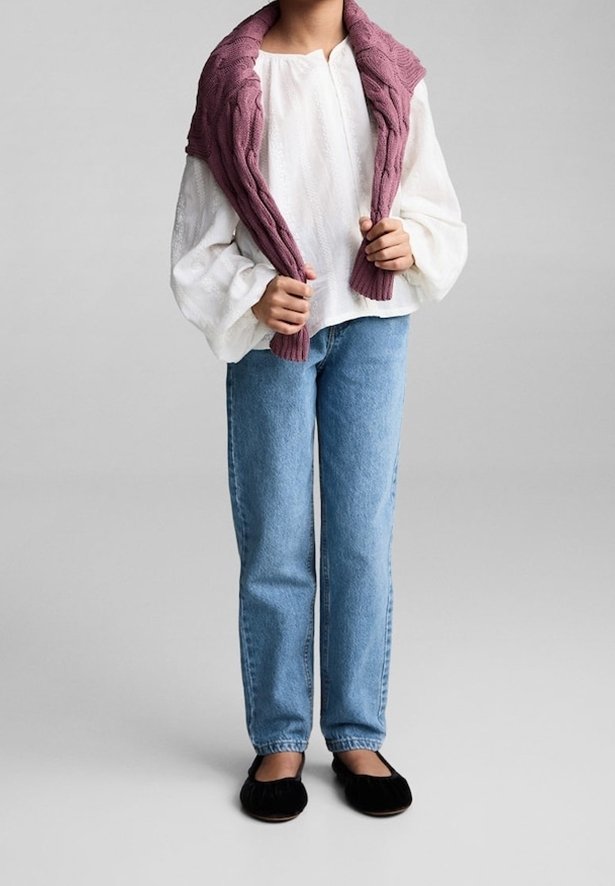 Personne portant un chemisier blanc, un jean bleu, des ballerines noires, tenant un pull en maille mauve drapé sur les épaules sur un fond gris.