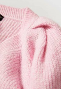 Světlerůžový pletený svetr s žebrovanou texturou a měkkým, chlupatým vzhledem. Má výstřih do V a detaily srolovaných rukávů.