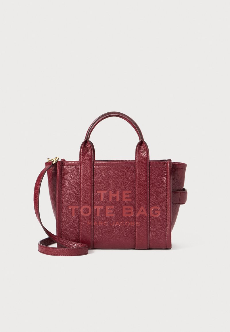 Saco tote em pele bordô com alças duplas e correia de ombro ajustável. Apresenta texto em relevé: "THE TOTE BAG MARC JACOBS." Superfície texturizada.