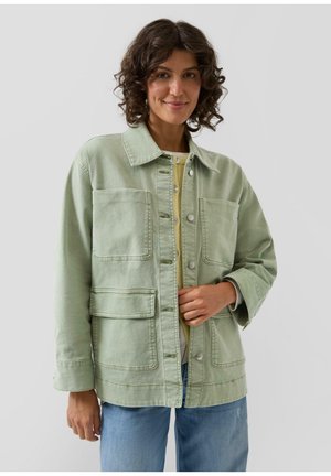 Vrouw met krullend haar, gekleed in een lichtgroene jasje met knopen over een geel topje en blauwe spijkerbroek, staand tegen een effen achtergrond.