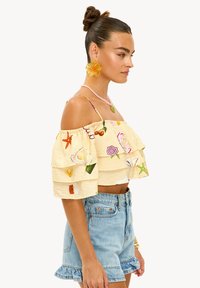 Haut jaune à épaules dénudées avec des couches à volants, orné de motifs brodés colorés ; associé à un short en denim bleu clair avec un ourlet effiloché.