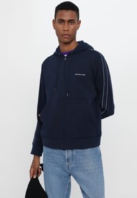 Felpa zip con cappuccio di colore blu navy, dotata di un cordino e rifinita con strisce laterali bianche e un piccolo logo "MICHAEL KORS" sul petto. Tessuto morbido.