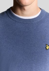 Maglione blu lavorato a maglia con colletto rotondo a coste e un piccolo logo giallo ricamato sul lato sinistro del petto. Tessuto morbido, design casual.
