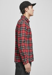 Rød og svart rutete skjorte med knappekrage, lange ermer og rett snitt. Brukt med en svart caps, vist i profil.