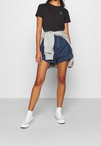 Camiseta de algodón negra con detalle gráfico verde, pantalones cortos de mezclilla azul de cintura alta, sudadera gris atada a la cintura y zapatillas altas blancas.