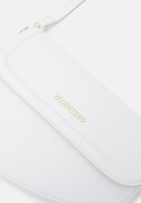 Bílá kožená kabelka přes rameno s klopovým uzávěrem, zdobená zlatým embosovaným logem "Valentino" a jemnými švy. Součástí je nastavitelné rameno.