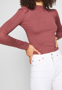 Haut à manches longues côtelé de couleur mauve avec des accents froncés aux épaules et un col montant, assorti à un jean en denim blanc avec des boutons métalliques visibles.