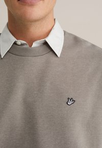 Sivá mikina s rebrovaným okrúhlym výstrihom, vrstvená cez bielu košeľu s golierom, s malým čierno-bielym vyšívaným logom vtáka.