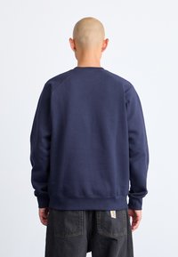 Marineblauw sweatshirt met een geribde onderrand en manchetten, achter raglanmouwen en een zachte textuur, gedragen met donkere broeken met een label op de achterzak.
