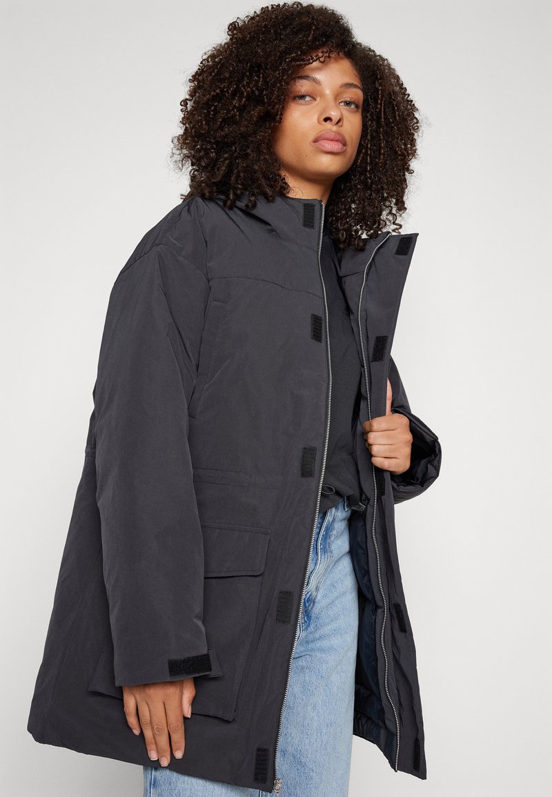 Veste sombre et isolée avec un col haut, fermeture éclair frontale et accents en Velcro. Dispose de poches latérales et d'une coupe ample.