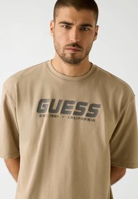 Beige crewneck t-shirt made of cotton, featuring a large gray "GUESS" logo and text. Short sleeves, relaxed fit, smooth texture.
