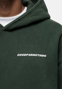 Ciemnozielona bluza z kapturem wykonana z mieszanki bawełny, z białym haftowanym napisem "GOODFORNOTHING" na wysokości klatki piersiowej oraz klasyczną kieszenią kangurką.