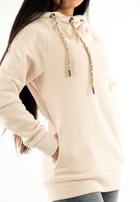 Sudadera rosa claro con capucha ajustable, que presenta cordones beige trenzados y puntas metálicas plateadas. Textura suave y puños y dobladillo acanalados.