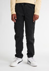 Zwarte cargobroek met elastische boorden, met zijzakken en een trekkoord in de taille, gedragen met witte sneakers en een lichtgele longsleeve top.