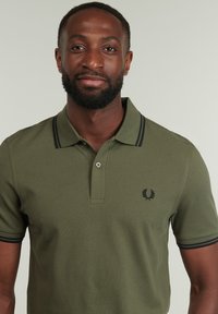 Polo de color verde oliva hecho de una tela texturizada, con franjas de acento negras en el cuello y un pequeño logotipo en el pecho.