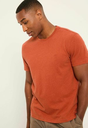 Mann trägt ein rostfarbenes kurzärmeliges T-Shirt, schaut nach unten und steht mit einer Hand in der Tasche einer beigen Hose vor einem neutralen Hintergrund.