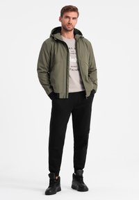 Giacca a zip verde oliva con cappuccio, abbinata a un maglione grigio chiaro e pantaloni neri. L'outfit è completato da scarpe alte nere.