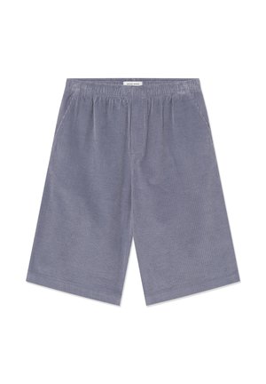 Mies, jolla on beige t-paita, harmaat ribilliset polvenmittaiset shortsit, valkoiset keskikorkeat sukat ja beiget urheilukengät, seisomassa valkoisella taustalla.
