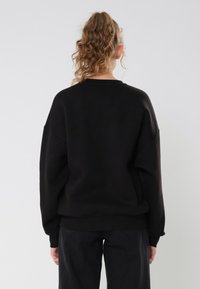 Černá oversized mikina s padajícími rameny, žebrovanými manžetami a lemem, vyrobená z měkké látky. Modelka je otočená zády, aby ukázala design na zadní straně.