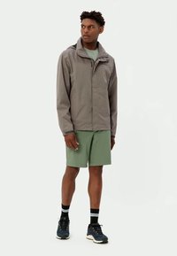 Vyras stovi apsirengęs pilka užtrauktuku striuke, žaliais šortais, juodomis kojinėmis su baltais dryžiais ir tamsiai mėlynais sportbačiais, žiūrėdamas į šoną.