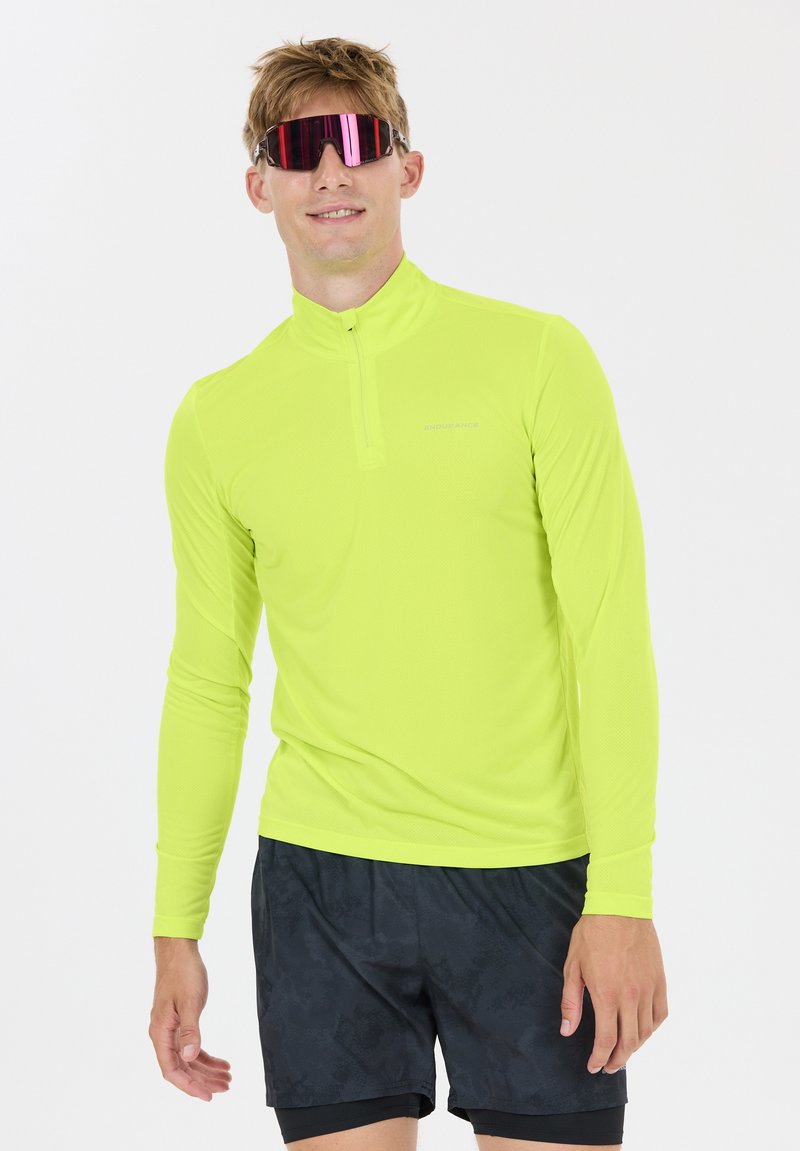 Homme portant un haut de sport à manches longues jaune néon et un short noir à motifs, avec des lunettes de soleil sportives réfléchissantes, debout.