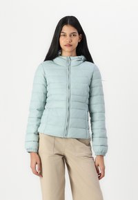 Lyseblå, polstret jakke med hette og glidelås. Har elastiske mansjetter og en figursydd silhuett. Bæres med beige bukser.