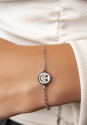 Pulsera de cadena de plata con un charm de cara sonriente, llevada en una muñeca, con fondo de camisa de manga larga blanca.