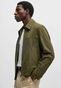 Olivgrön mockajacka med en modern krage, dragkedjestängning och sidofickor, bärs över en vit skjorta och beiga byxor. Slät textur.