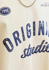 Kokvilnas T-krekls krēmkrāsā ar tumši zilu drukātu tekstu "ORIGINAL studio" un "1990". Ievērojama ir gluda tekstūra un neliela spīduma klātbūtne.