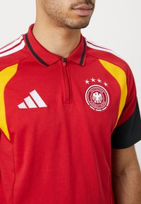 Κόκκινο πόλο μπλουζάκι με μαύρο γιακά και φερμουάρ, διαθέτει κίτρινες ένθετες παλέτες, λευκό λογότυπο Adidas και κεντημένο έμβλημα της γερμανικής ποδοσφαιρικής ομάδας με αστέρια.