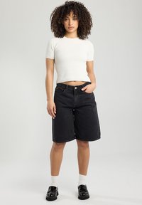 Black denim bermuda shorts with a loose fit, high waist, and rolled hems, paired with a fitted white ribbed short-sleeve top. Black shoes and white socks.