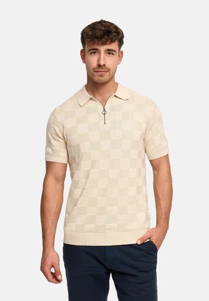 Mężczyzna ubrany w beżową, krótkim rękawem, dzianinową koszulkę polo z suwakiem do ćwiartki i wzorem w kratę, w połączeniu z granatowymi spodniami, stojący na białym tle.