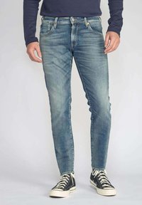 Jeans en denim bleu délavé avec une coupe slim, un design à cinq poches et des coutures contrastées, associés à des baskets montantes noires.