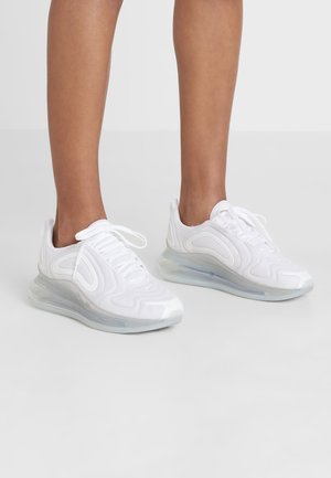 Ben iført hvide sports-sneakers med tykke, gennemsigtige polstrede såler på en ensartet lysegrå baggrund.